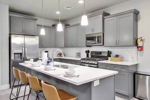 A modern kitchen with grey cabinets, a central island with bar stools, stainless steel appliances, and pendant lighting.