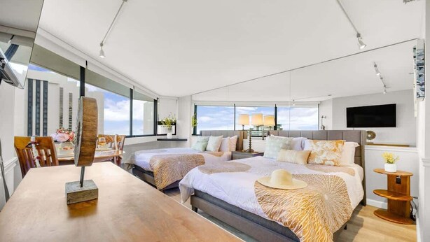 A modern hotel room with two beds, a wooden dining table, and a large window offering a view of the city skyline. - Waikiki Forever Ocean Views, Free Parking & Pool (Honolulu)