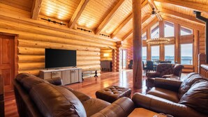 Een houten blokhut inrichting met een groot houten plafond, een televisie, leren banken en een eethoek met uitzicht op de natuur.