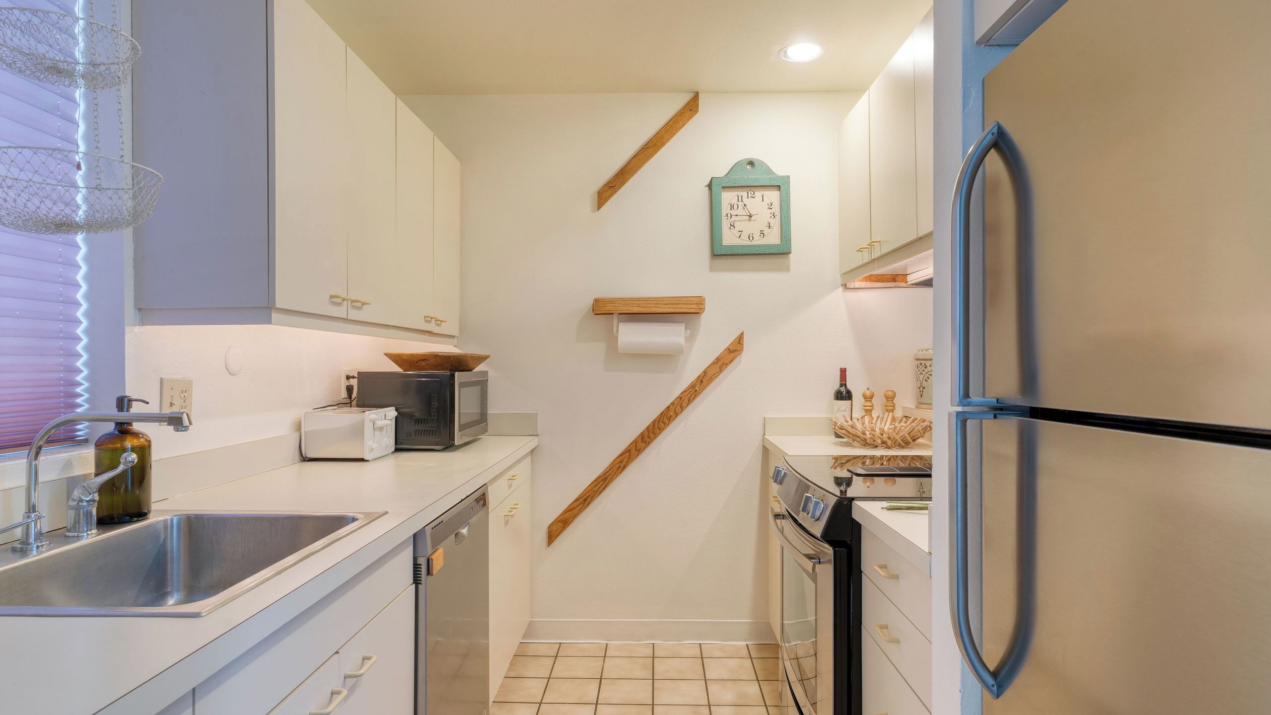 A kitchen with a sink, toaster, microwave, wooden shelf, and a clock on the wall.