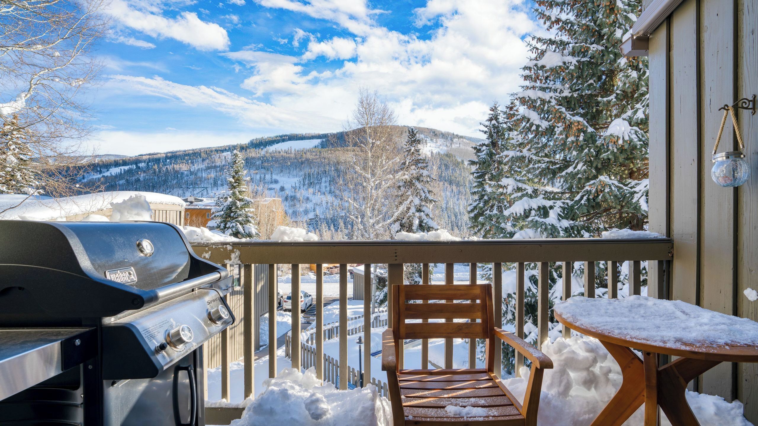 A balcony with a grill, a wooden chair, and a table covered in snow, overlooking a snowy mountain landscape.