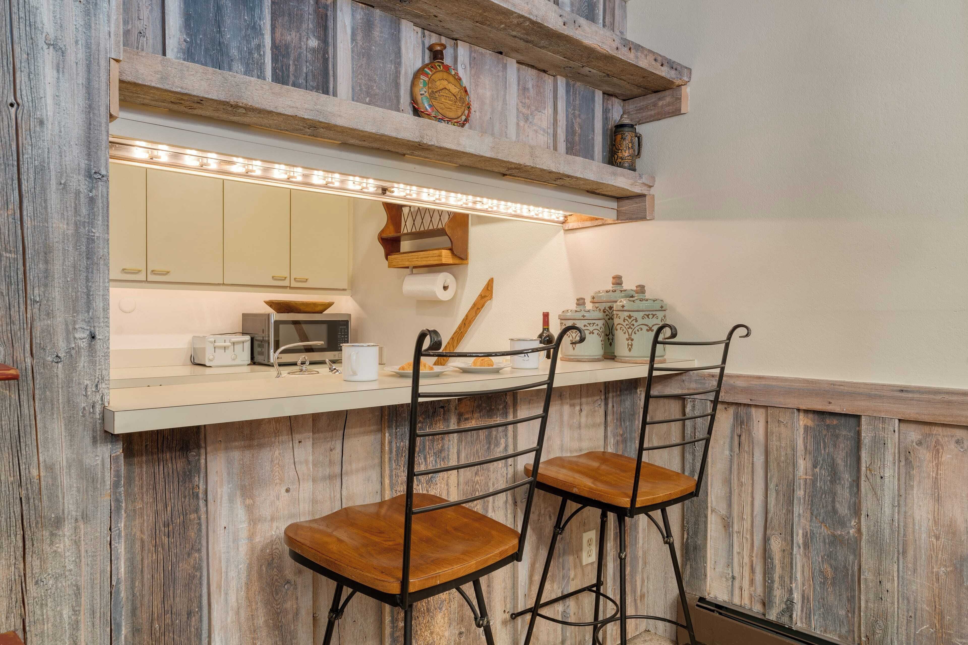 A rustic kitchen with wooden walls, a white countertop, two bar stools, and a hanging light fixture.