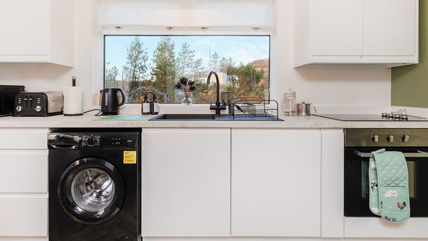 Cucina moderna con lavatrice integrata, lavello e finestra vista alberi e colline.