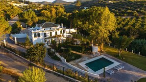 A large white villa with a swimming pool, surrounded by lush greenery and hills in the background.