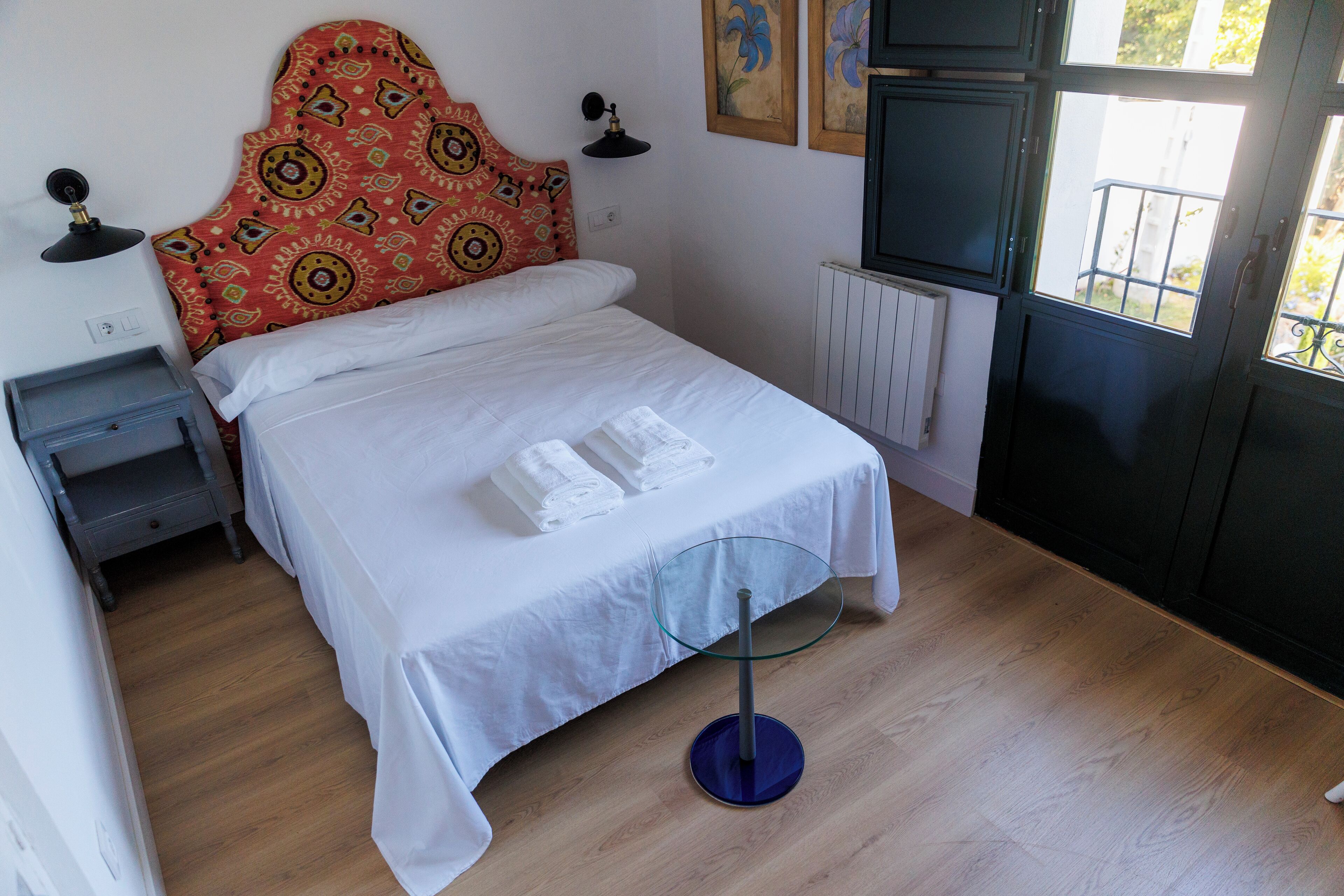 Un lit bien fait, avec du linge de lit blanc, une table de chevet en verre, une tête de lit à motifs et une vue sur l’extérieur par une fenêtre.