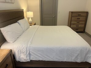 Een keurig opgemaakt bed met witte lakens, een houten hoofdbord, een bedlampje en een houten dressoir met lades.