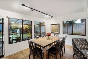 A dining area with a wooden table and chairs, large windows, and artwork on the walls.