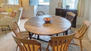 Un coin repas avec une table ronde en bois, six chaises en bois, un saladier de fruits, un canapé avec des coussins à motifs floraux, une lampe et une vue sur l’extérieur par une fenêtre.
