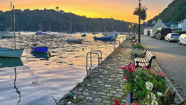 Une promenade en bord de mer avec des bateaux amarrés, un chemin pavé, un banc et une rue bordée d’immeubles.