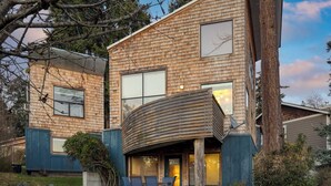 A two-story wooden house with a curved roof over a patio area, surrounded by a small garden and trees. - Deck Views, Fire Pit, Pool, 3 Mins to Beach Madrona Tree Retreat by Avantstay (Coupeville)