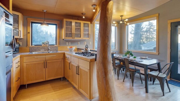 A modern kitchen with wooden cabinets, a central island, and a dining area with chairs and a table. - Deck Views, Fire Pit, Pool, 3 Mins to Beach Madrona Tree Retreat by Avantstay (Coupeville)
