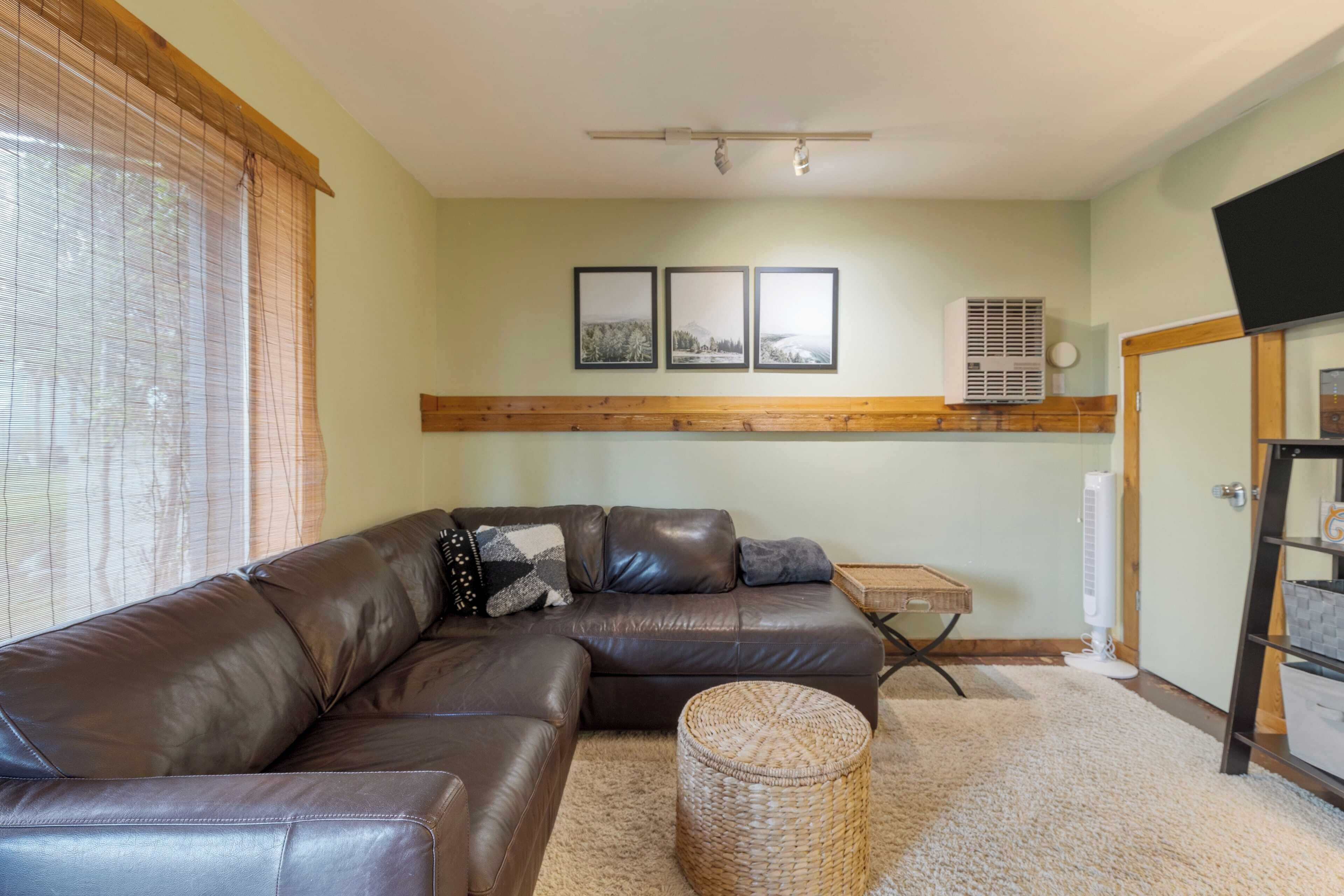 A living room with a brown leather sectional sofa, a wicker ottoman, and framed pictures on the wall.