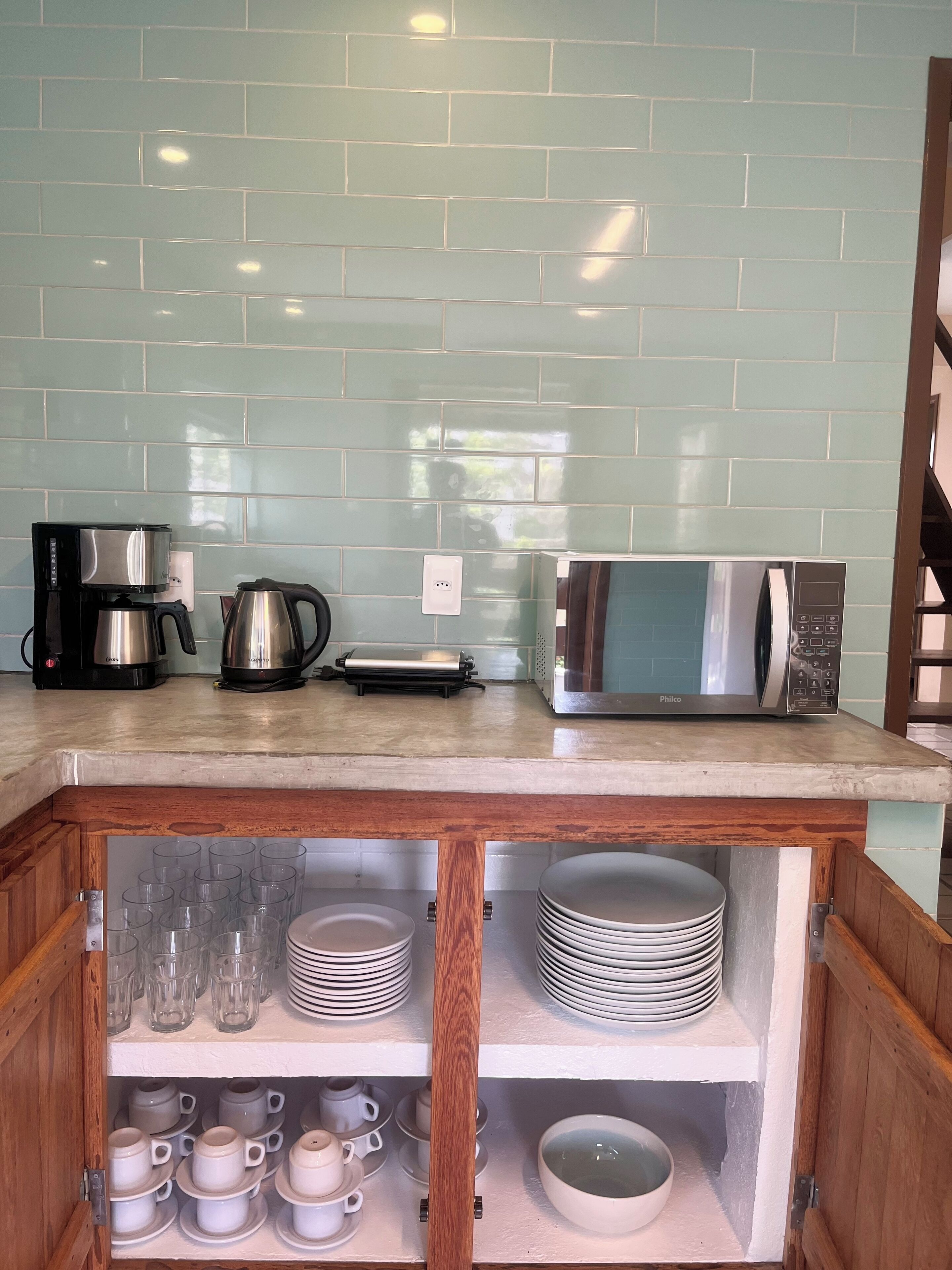Cozinha com painel de azulejos, cafeteira, chaleira, micro-ondas e armário de madeira com pratos e copos.