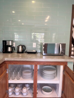 Cozinha com painel de azulejos, cafeteira, chaleira, micro-ondas e armário de madeira com pratos e copos.