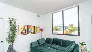 Una sala de estar moderna con un sofá seccional verde, una mesa de centro de vidrio y un ventanal que ofrece vistas a palmeras y edificios.