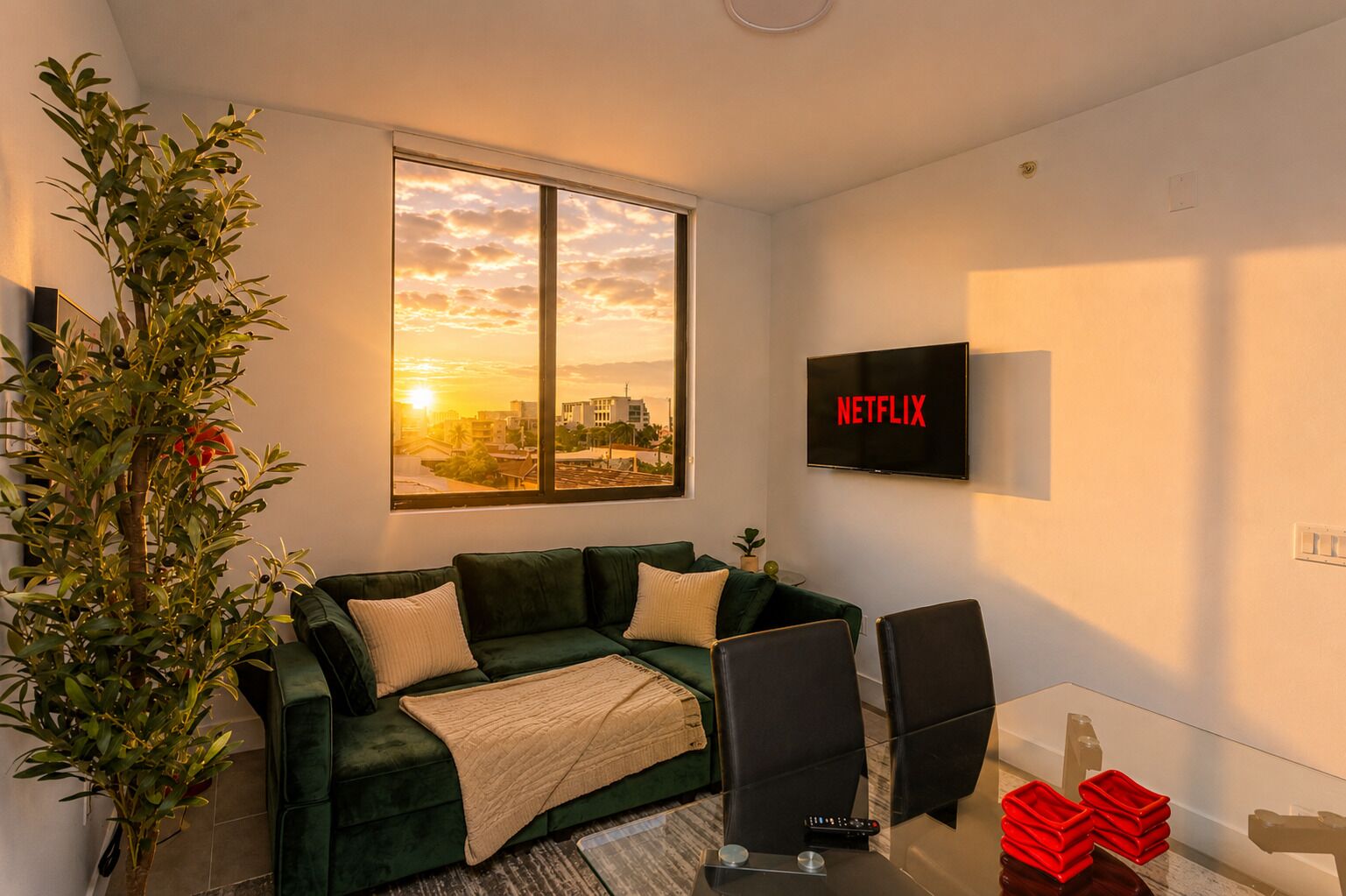 A living room with a green sofa, a TV mounted on the wall, and a large window with a sunset view.