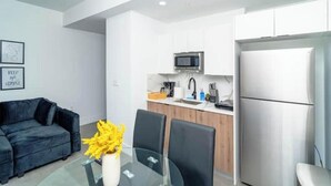 Una cocina moderna con armarios blancos, un microondas y un refrigerador. Hay un comedor con una mesa de vidrio y sillas negras.