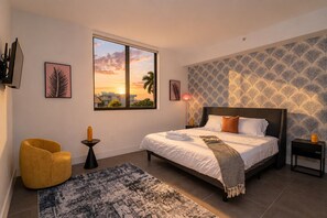 A bedroom with a bed, a yellow chair, a nightstand, a lamp, and a large window with a sunset view.