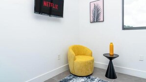Una habitación moderna con un sillón amarillo, una mesita negra pequeña con una botella amarilla, una obra de arte con una hoja enmarcada y un televisor mostrando 'NETFLIX'.