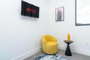 A modern room with a yellow armchair, a small black table with a yellow bottle, a framed leaf artwork, and a TV displaying 'NETFLIX'.