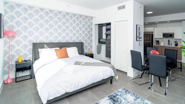 A modern bedroom with a large bed, bedside tables, a desk with chairs, and a kitchenette in the background.
