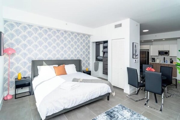 A modern bedroom with a large bed, bedside tables, a desk with chairs, and a kitchenette in the background.