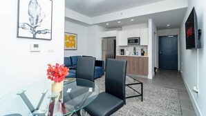 Una sala de estar moderna con una mesa de centro de vidrio, sillas negras, un sofá azul y una zona de cocina con armarios blancos y un microondas.