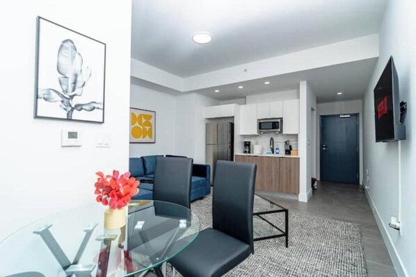 Una sala de estar moderna con una mesa de centro de vidrio, sillas negras, un sofá azul y una zona de cocina con armarios blancos y un microondas.