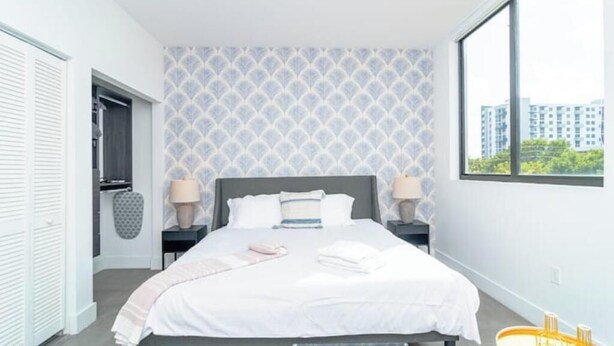 A modern bedroom with a large bed, two bedside tables, a window with a city view, and a patterned wall. - King Bed brand new safe Oasis minutes to Brickell (Miami)
