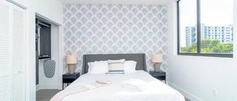 A modern bedroom with a large bed, two bedside tables, a window with a city view, and a patterned wall.