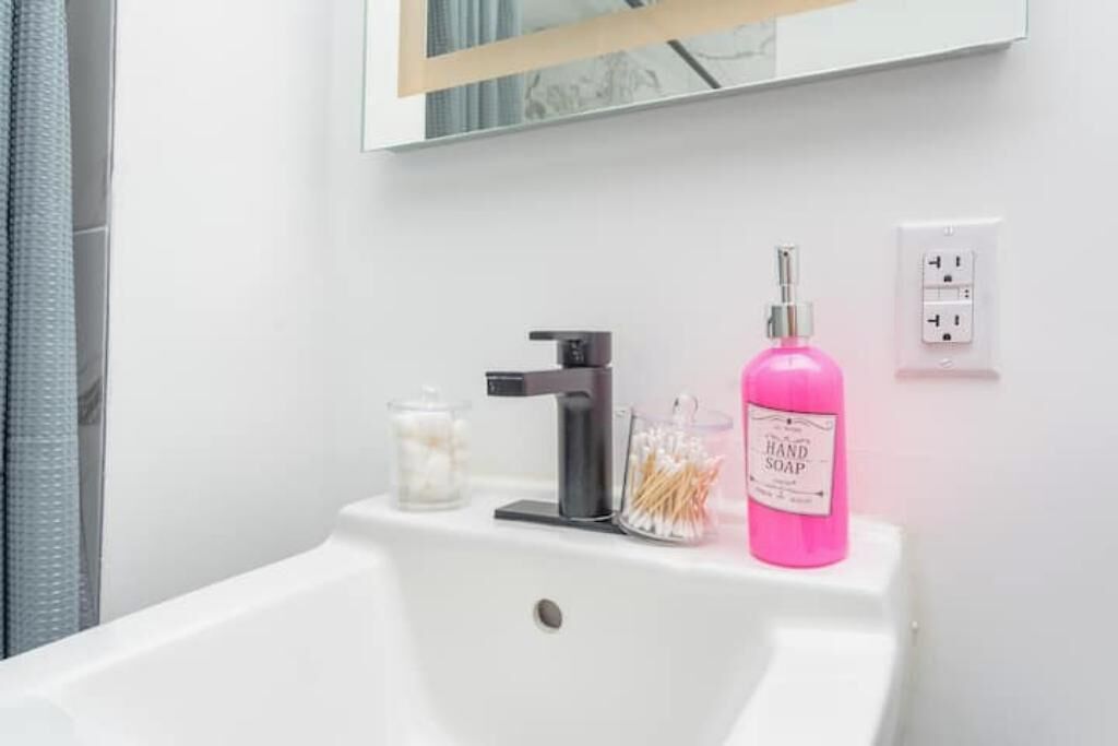 Uma pia de banheiro com uma torneira, um dosador de sabonete rosa e um recipiente de vidro com cotonetes.