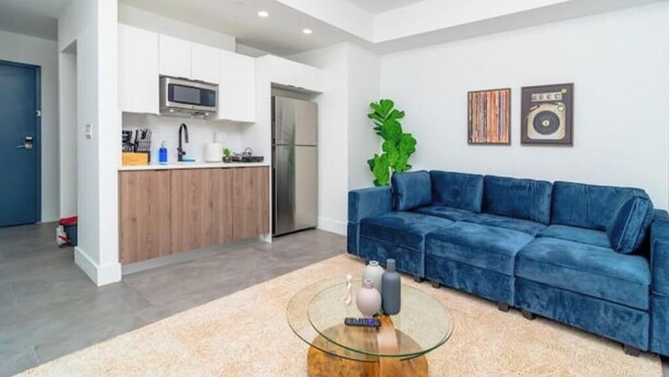 Una sala de estar moderna con un sofá azul, una mesa de centro de vidrio y una zona de cocina con armarios blancos y un microondas.