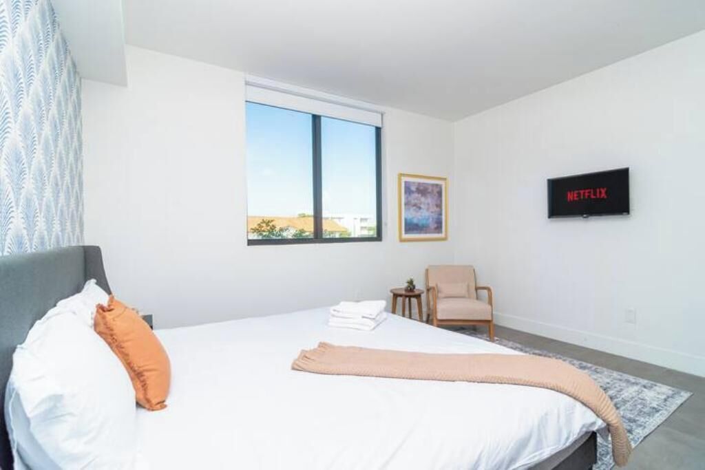 Une chambre à coucher avec un lit, une chaise, une petite table, une fenêtre et un téléviseur fixé au mur.