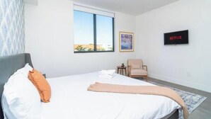 Un dormitorio con cama, una silla, una mesita, una ventana y un televisor en la pared.