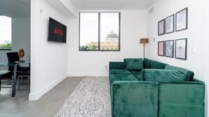 Una sala de estar moderna con un sofá verde, un televisor de pantalla plana montado en la pared y un ventanal con vistas a un edificio.