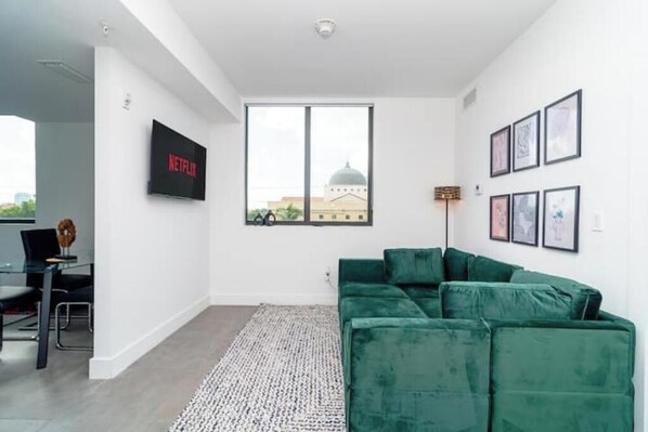 A modern living room with a green sofa, a flat-screen TV mounted on the wall, and a large window with a view of a building. - Miami Semi 2BR Getaway with King Beds Near Marlins Park & Calle Ocho (Miami)