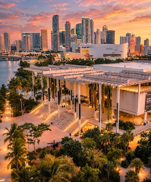 A modern building with a rooftop terrace, palm trees, and a city skyline in the background.