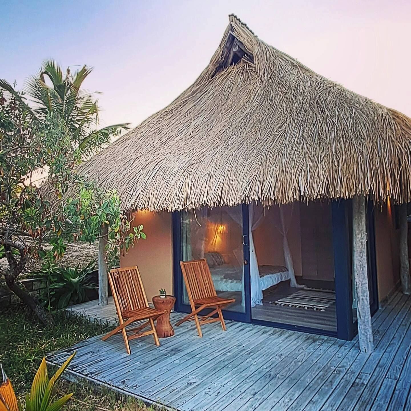 A thatched-roof bungalow with two wooden chairs and a small table on a wooden deck.