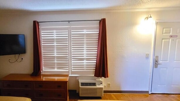 Une pièce avec un téléviseur fixé au mur, une fenêtre avec des stores, un radiateur, une commode en bois et une porte.