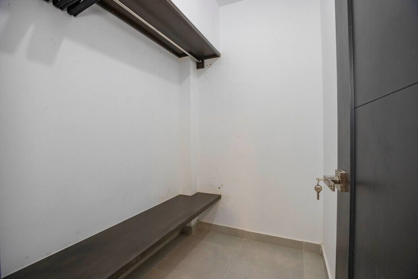 Une petite pièce vide, avec un banc en bois sombre, une étagère en métal et une porte dotée d’une serrure à clé.