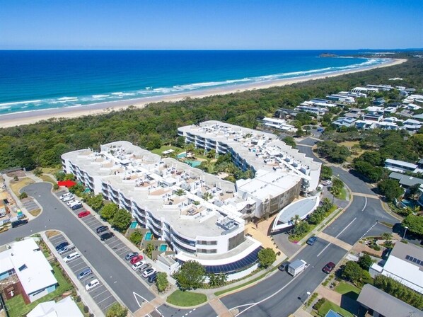 An aerial view of a coastal hotel with multiple buildings, parking areas, and a beach in the background.