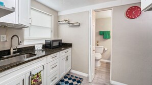 Une petite cuisine équipée d’un évier, d’un four à micro-ondes et de placards, donnant sur une salle de bain avec des toilettes et une horloge au mur.