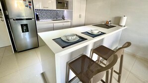 A modern kitchen with white cabinetry, a stainless steel refrigerator, a microwave, and a gas stove. There are two chairs and a small island with a sink and a bowl. - Your Home Away: Rooftop Pool Close to Beach & Fun (Playa del Carmen)