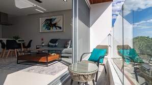 A modern living room with a sofa, coffee table, and chairs, a dining area with chairs and a table, and a balcony with wicker chairs and a glass railing. - Your Home Away: Rooftop Pool Close to Beach & Fun (Playa del Carmen)