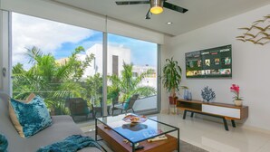 A modern living room with a glass coffee table, a sofa with a blue throw blanket, a TV mounted on the wall, and a balcony with plants. - Your Home Away: Rooftop Pool Close to Beach & Fun (Playa del Carmen)