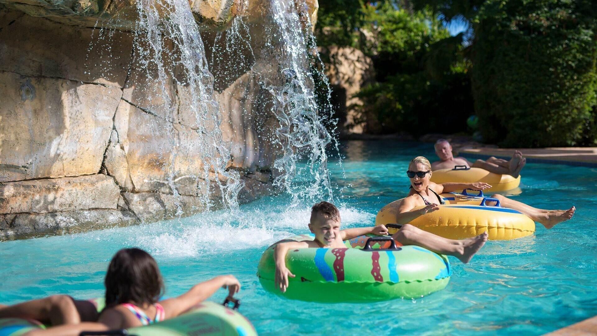 A pool with people floating on inflatable rings, a waterfall feature, and a rocky backdrop.