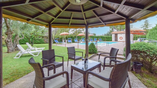 Un espace extérieur aménagé, abrité et doté de chaises en osier et d’une table, offrant une vue sur une piscine et des chaises longues.