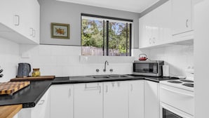 Una cucina moderna con mobili bianchi, un piano di lavoro nero, un forno a microonde, un bollitore e una finestra con vista sugli alberi.