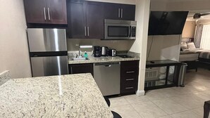 A compact kitchen with dark cabinets, stainless steel appliances, a granite countertop, and a small dining area with a chair.