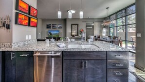 A modern kitchen with granite countertops, stainless steel appliances, and a large island. There are pendant lights, a dining area with a table and chairs, and large windows offering a view. - This Is It -Gorgeous Southwest Modern Luxury! (Alto)
